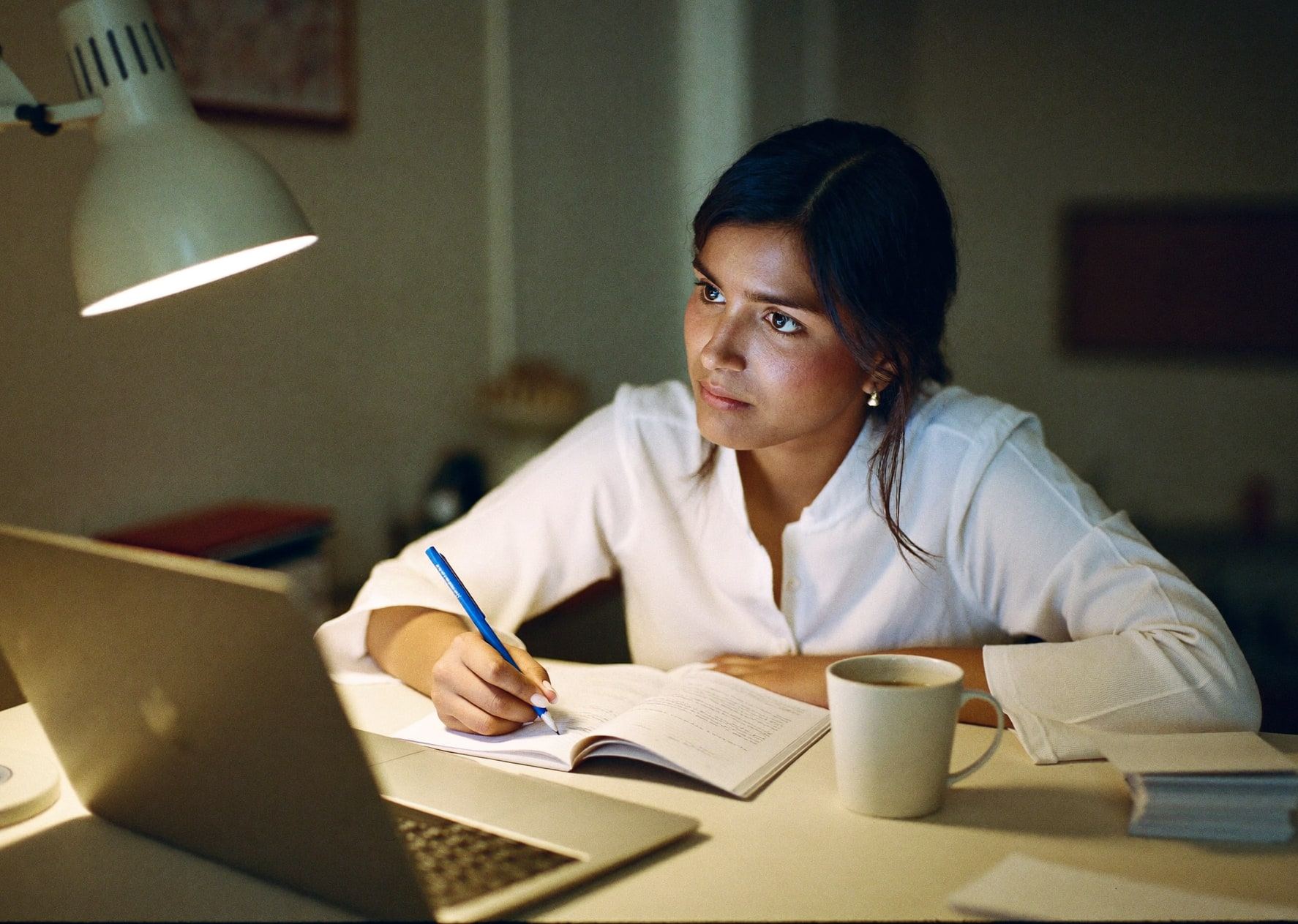 A woman studying at a home desk at night by warm lamplight