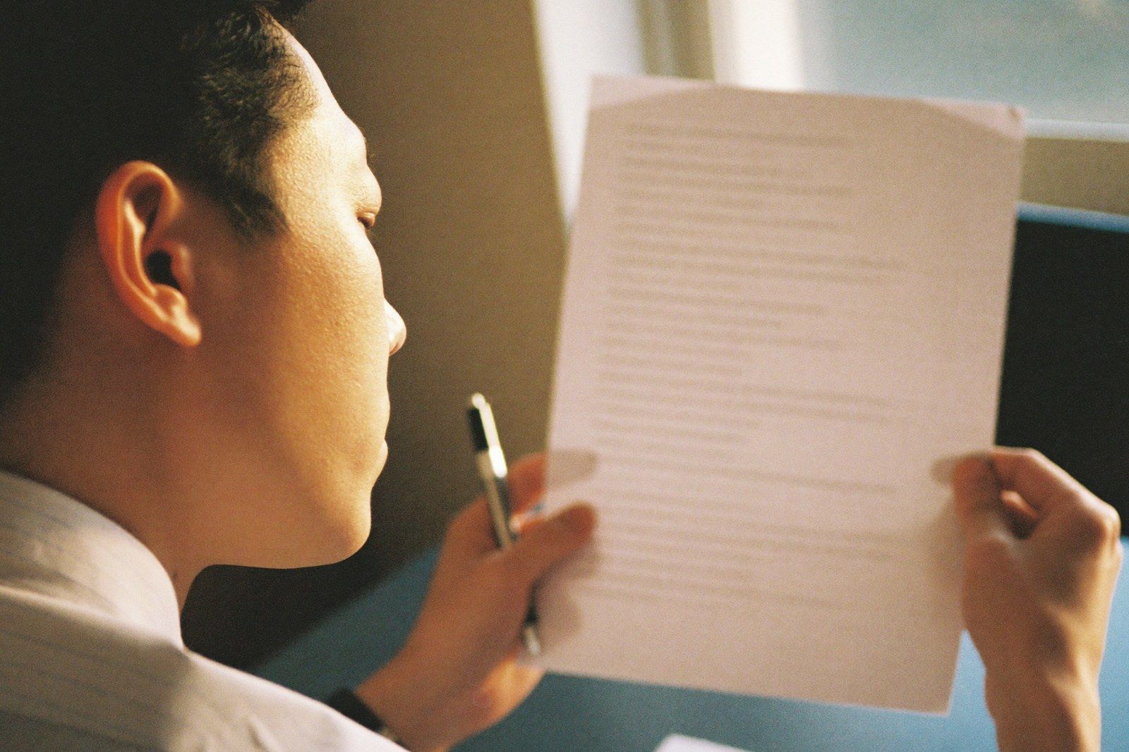 A person seen over the shoulder reading a printed page of text closely, a pen held ready in one hand, by a bright window.
