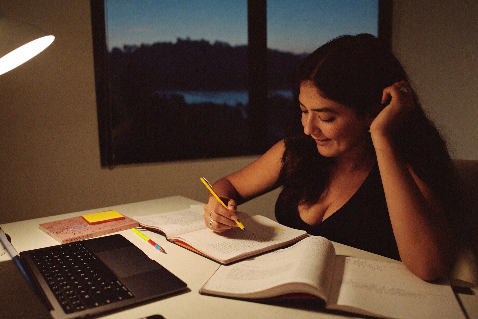 A woman writing in a notebook at a desk in the evening, an open textbook and a laptop in front of her and a lamp lighting the page.