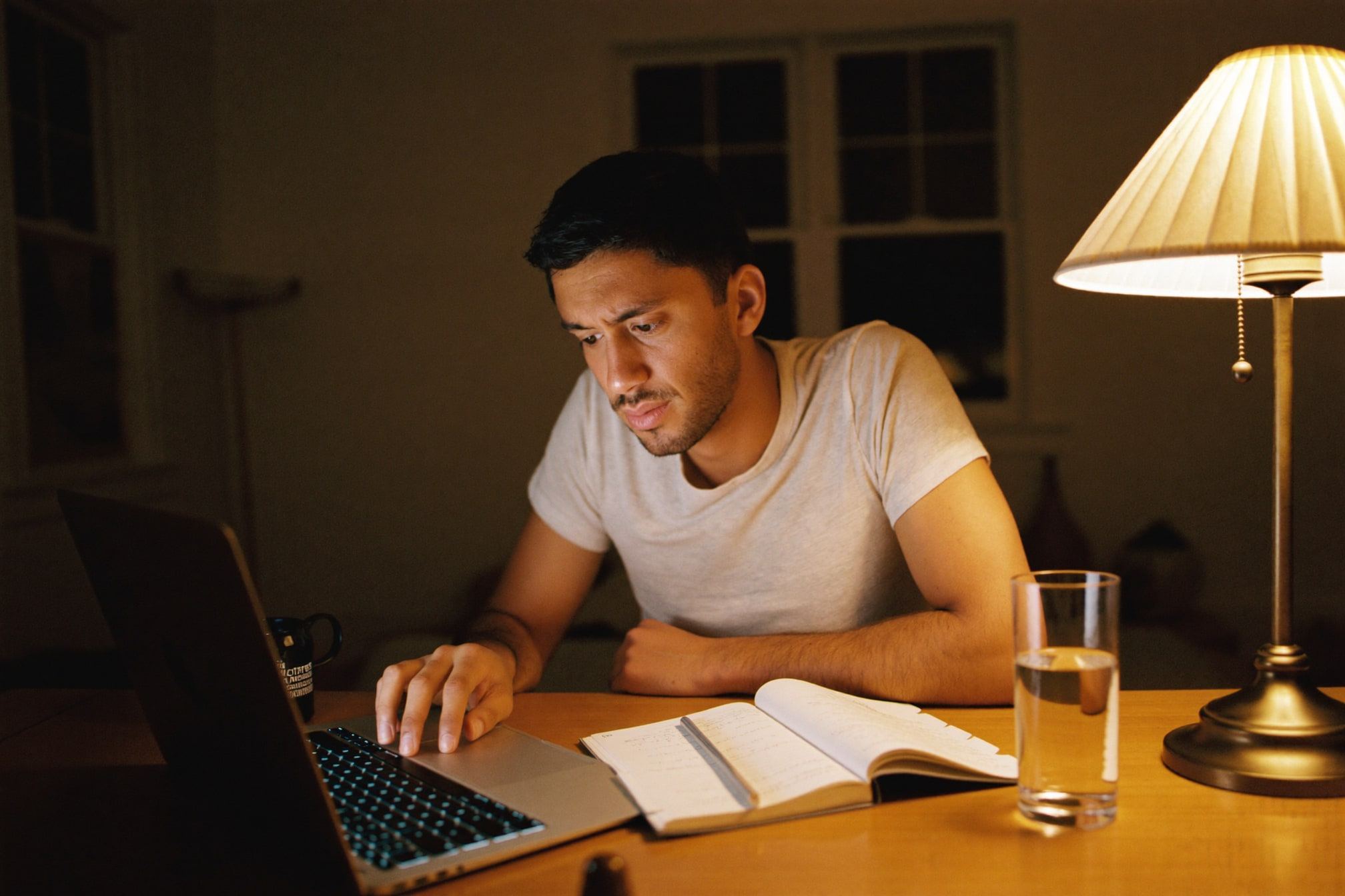 A working parent studying from a notebook at the dining table late at night, lit by a single warm lamp
